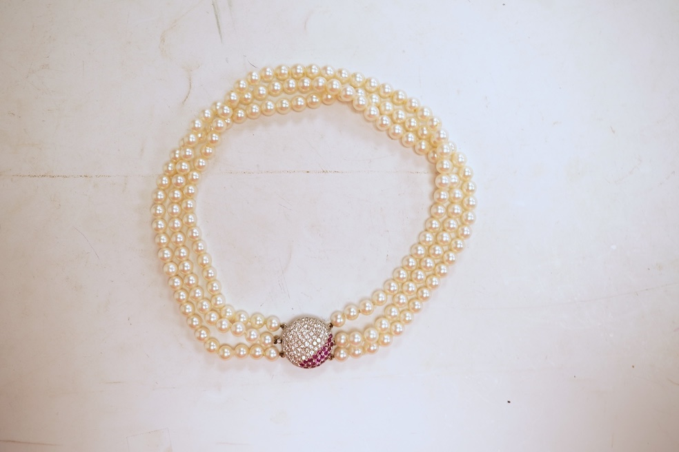 A cultured pearl, ruby and diamond clasp, composed of three rows of cultured pearls measuring approximately 6.9mm diameter, to a white precious metal (tests as 14k) disc-shaped clasp pavé-set with brilliant-cut diamonds